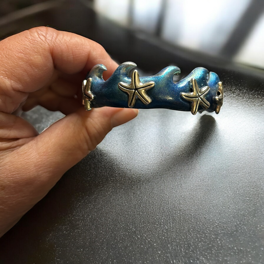 Decorative bracelet with starfish and wave design held by a hand on a dark surface.