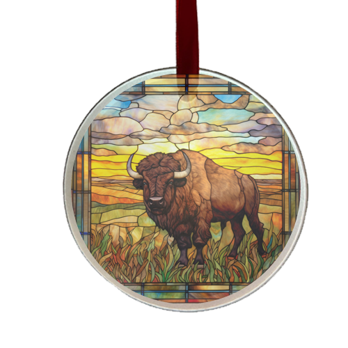A round glass ornament featuring a stained glass design with a bison in the foreground, set against a sunset-like background, and a satin ribbon at the top.