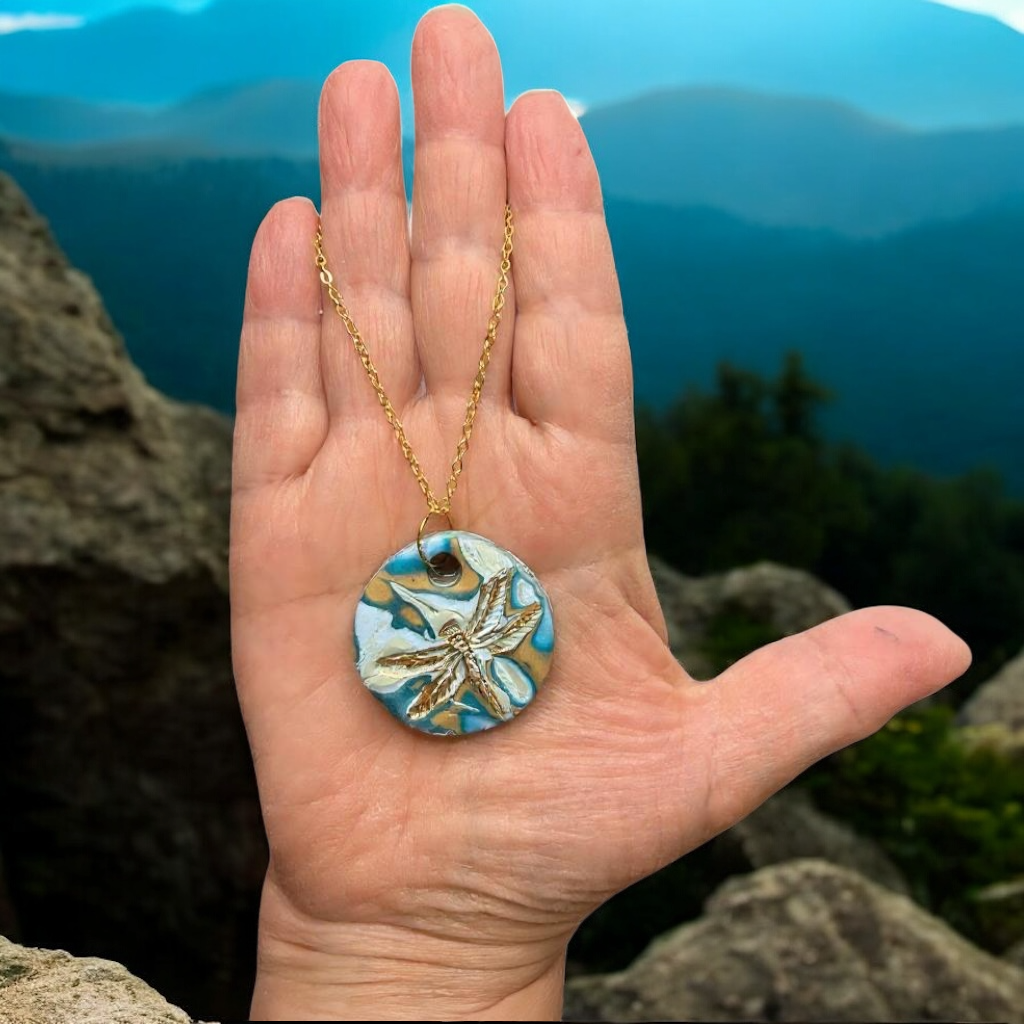 Hand holding a necklace with a dragonfly pendant against a mountainous background