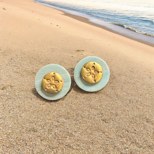Gold sand dollar earrings on a sandy beach with ocean in the background