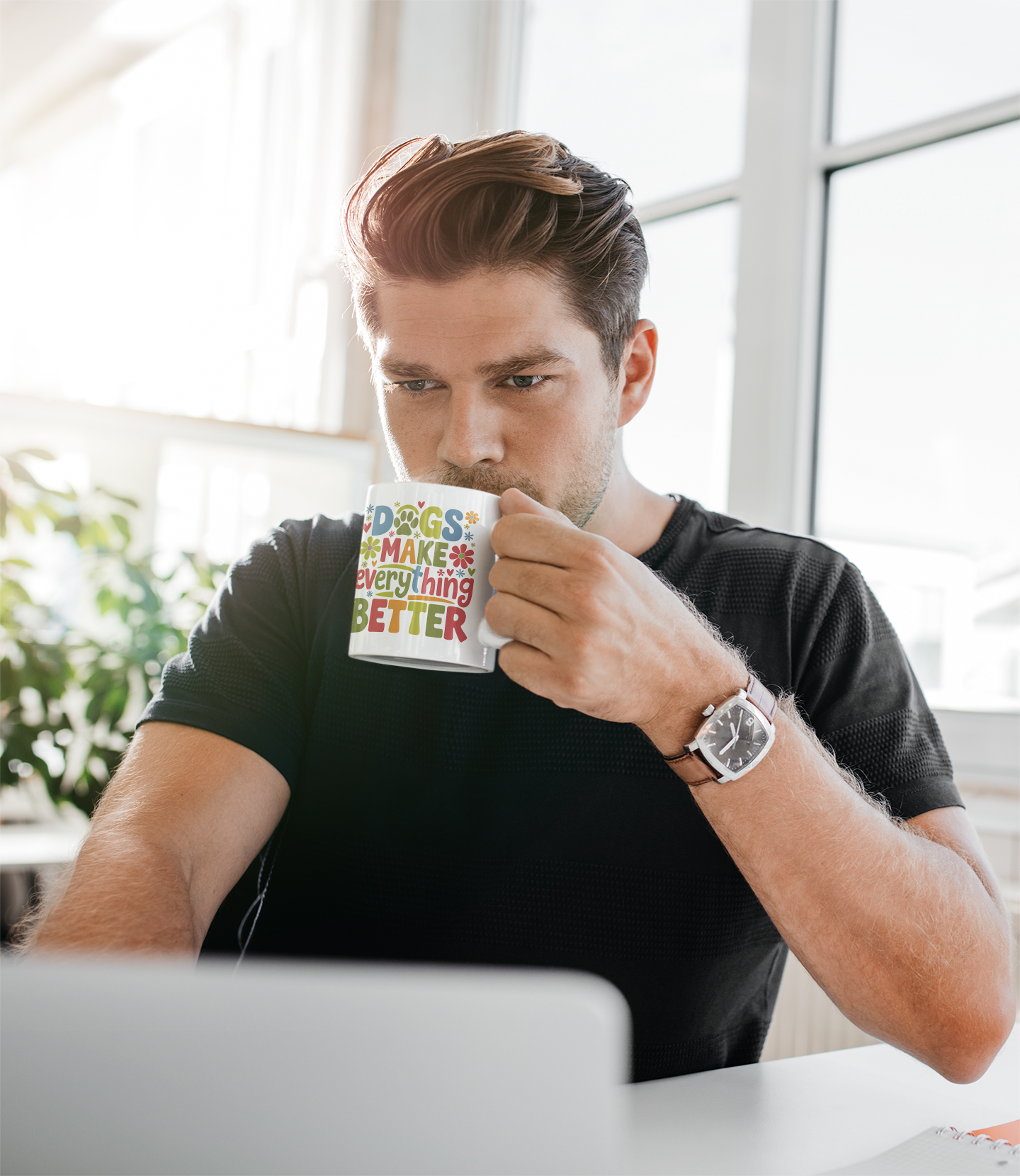 Man drinking from a mug with text, "Dos make everything better", sitting at a desk in a bright room.